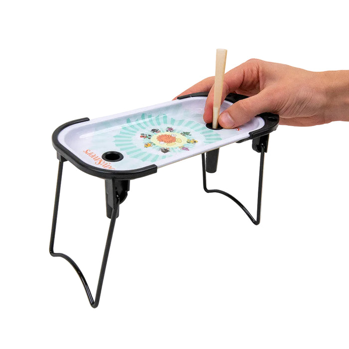 A hand holding a wooden stick over a small tabletop game tray with a colorful printed center and black metal legs, shown on a clean white background.